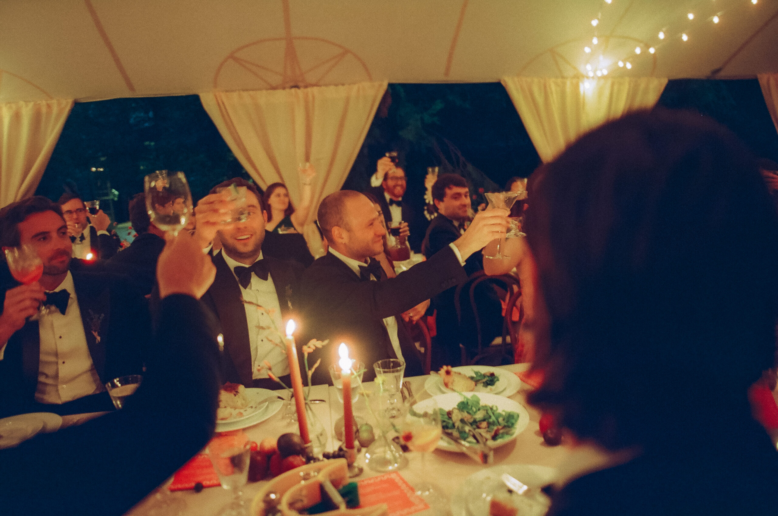 At a tented reception, guests hold up their glasses for a toast cheers with the grooms centered, arms around one another.